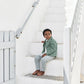 A young boy sits barefoot on white outdoor steps, wearing the Tartine et Chocolat Baby Boys’ and Boys’ Explorer Jacket with striped pants, gazing slightly to his left. White walls and a railing frame the scene.