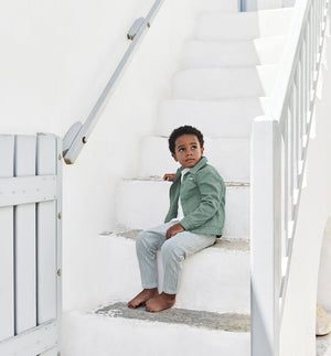 A young boy sits barefoot on white outdoor steps, wearing the Tartine et Chocolat Baby Boys’ and Boys’ Explorer Jacket with striped pants, gazing slightly to his left. White walls and a railing frame the scene.
