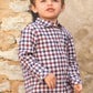 A young child with light brown hair, wearing the Tartine et Chocolat Baby Boys’ and Boys’ Cotton Check Shirt and beige pants, stands against a stone wall and looks slightly upward.