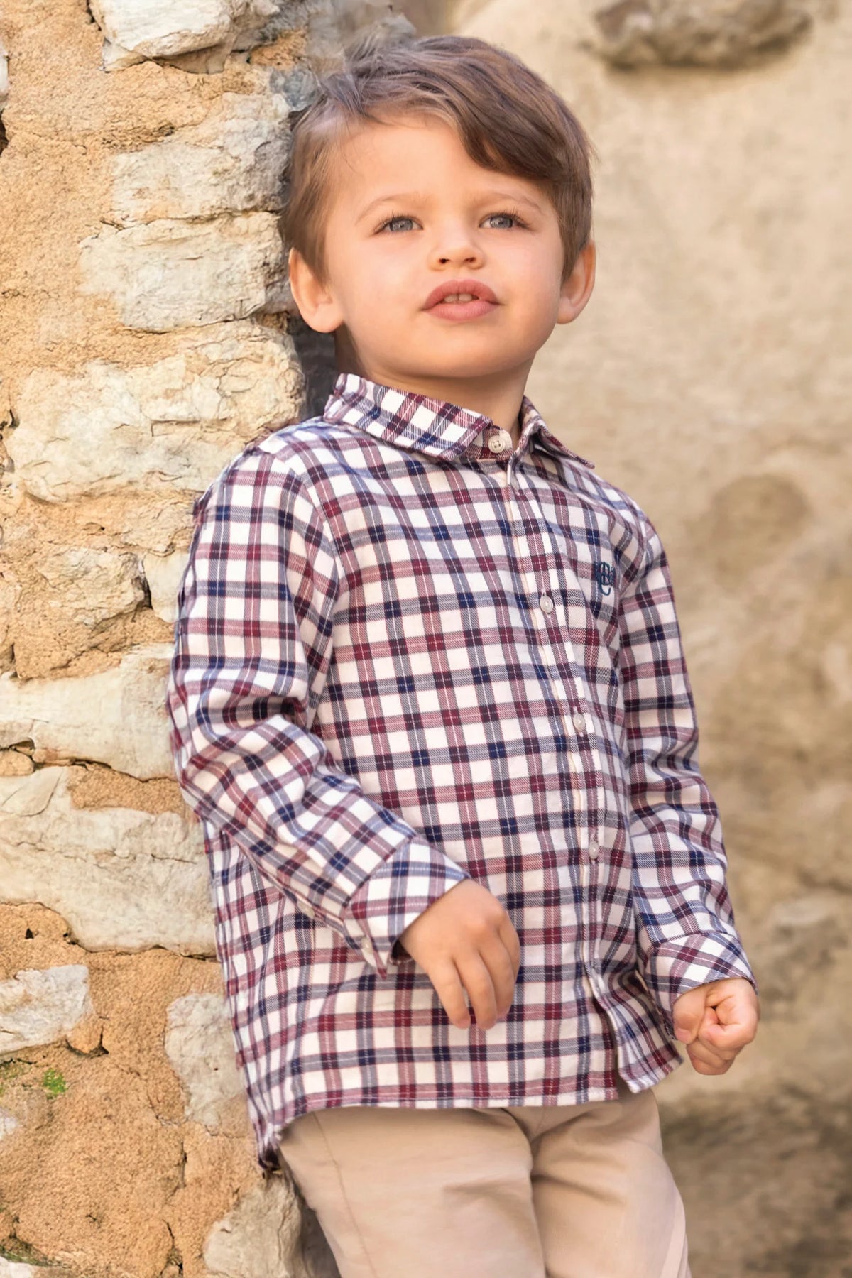 A young child with light brown hair, wearing the Tartine et Chocolat Baby Boys’ and Boys’ Cotton Check Shirt and beige pants, stands against a stone wall and looks slightly upward.