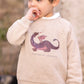 A young child stands outdoors, sucking their thumb, dressed in the Tartine et Chocolat Baby Boys’ and Boys’ Dinosaur Print Cotton Sweater.