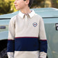 A boy wearing the Tartine et Chocolat Boys’ Pointed Collar Stripes Polo Shirt, featuring beige, navy, and burgundy cotton with an embroidered crest, stands outdoors next to a light blue vehicle and gazes to the side—a scene that captures timeless style.