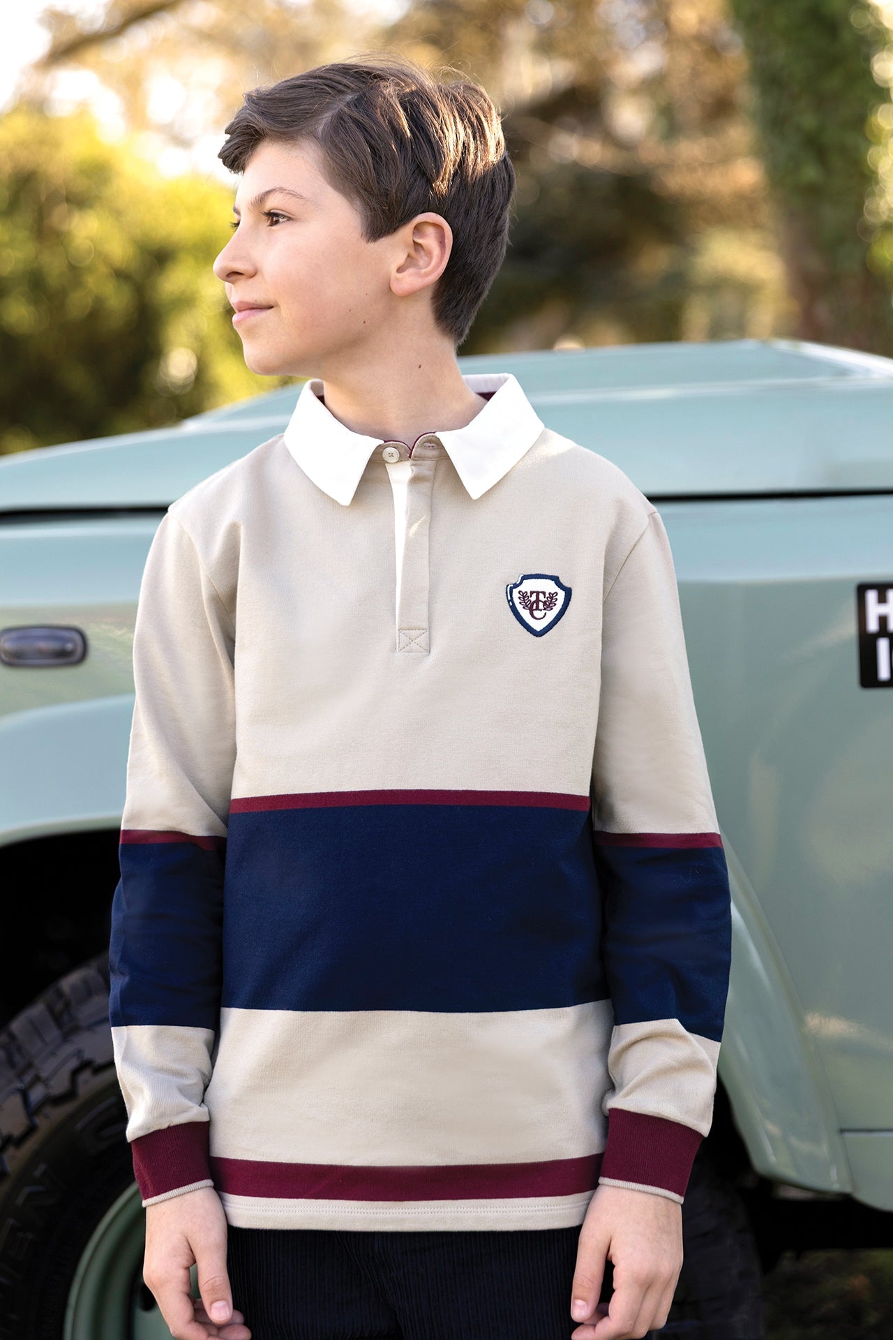 A boy wearing the Tartine et Chocolat Boys’ Pointed Collar Stripes Polo Shirt, featuring beige, navy, and burgundy cotton with an embroidered crest, stands outdoors next to a light blue vehicle and gazes to the side—a scene that captures timeless style.