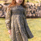 A young girl with long red hair is wearing the Tartine et Chocolat Girls’ Liberty Embroidery Round Neck Cotton Dress and black boots as she stands on grass in front of a stone wall and building, gazing slightly to the side.