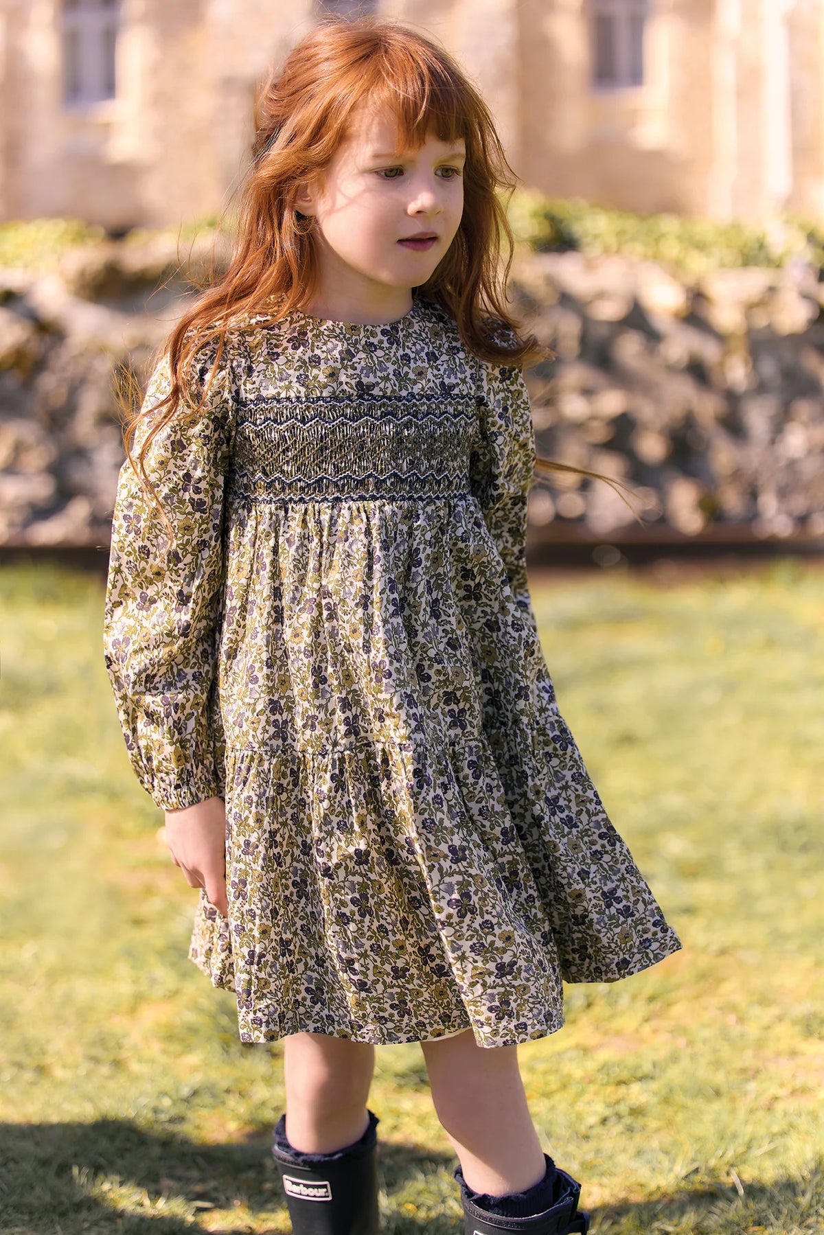 A young girl with long red hair is wearing the Tartine et Chocolat Girls’ Liberty Embroidery Round Neck Cotton Dress and black boots as she stands on grass in front of a stone wall and building, gazing slightly to the side.