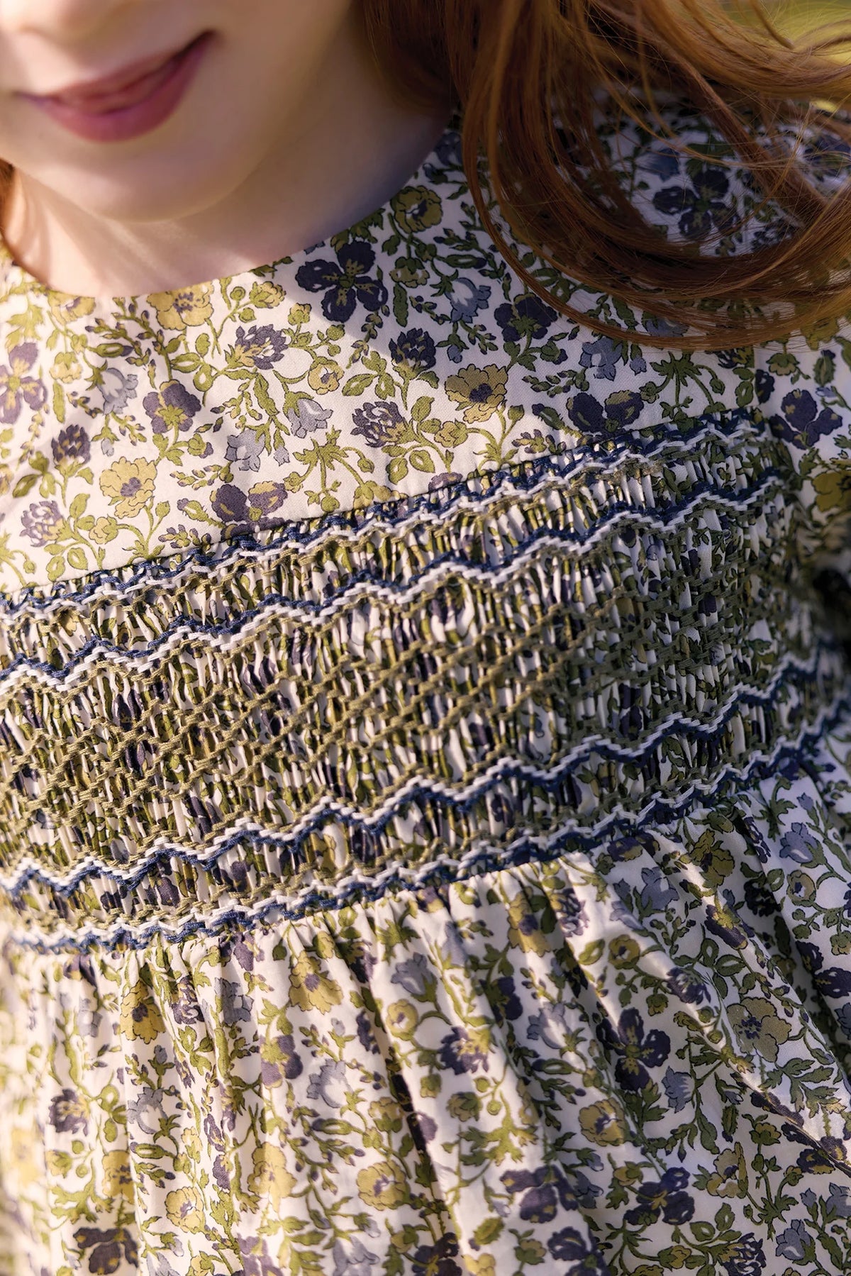 Close-up of a child wearing the Tartine et Chocolat Girls’ Liberty Embroidery Round Neck Cotton Dress in olive cotton, featuring intricate smocking across the chest and delicate Liberty floral embroidery; partial view of face and brown hair evokes the charm of an English garden.