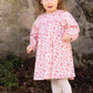 A young child wearing the Tartine et Chocolat Girls’ Floral Print Cotton Dress, white tights, and gold shoes stands outdoors near a stone wall and some greenery.