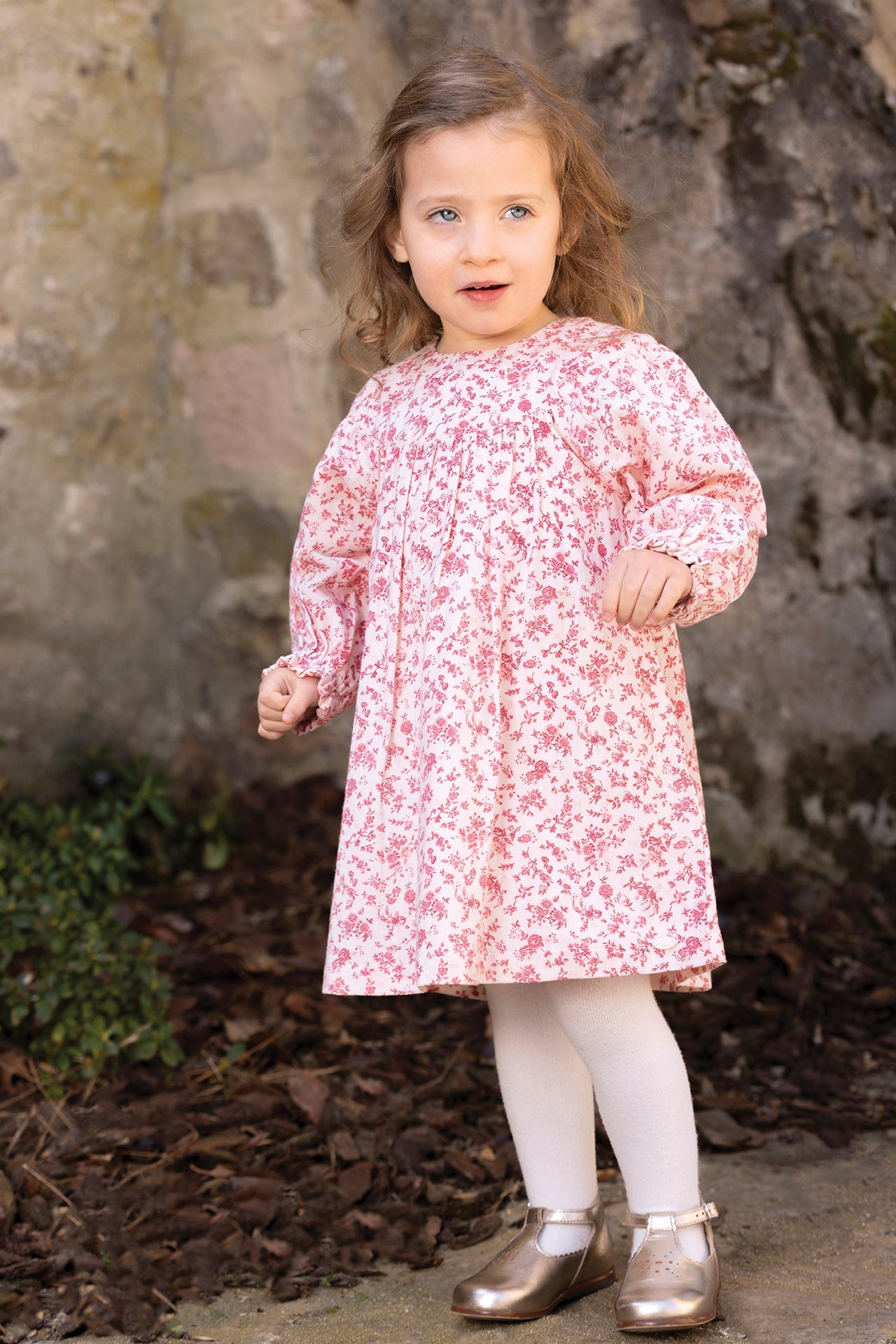A young child wearing the Tartine et Chocolat Girls’ Floral Print Cotton Dress, white tights, and gold shoes stands outdoors near a stone wall and some greenery.