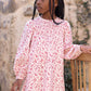 A young girl with braided hair, dressed in the Tartine et Chocolat Girls’ Flowery Print Cotton Dress with long sleeves and a smocked waist, stands beside a wooden chair and gazes to the side. A stone wall and climbing plants create a charming backdrop.