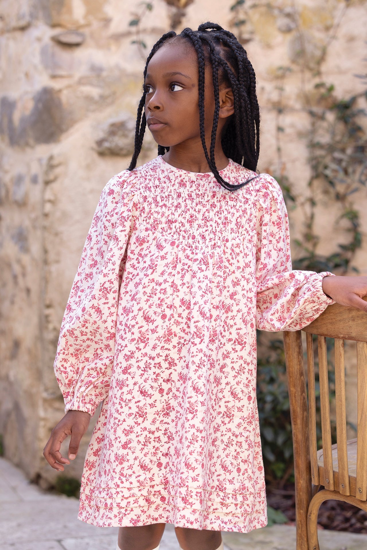 A young girl with braided hair, dressed in the Tartine et Chocolat Girls’ Flowery Print Cotton Dress with long sleeves and a smocked waist, stands beside a wooden chair and gazes to the side. A stone wall and climbing plants create a charming backdrop.