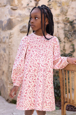 A young girl with braided hair, dressed in the Tartine et Chocolat Girls’ Flowery Print Cotton Dress with long sleeves and a smocked waist, stands beside a wooden chair and gazes to the side. A stone wall and climbing plants create a charming backdrop.
