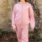 A young girl wearing the Tartine et Chocolat Girls’ Wool Hood Cotton Jogging Suit, featuring a zip-up hoodie and matching pants in pink, stands outdoors on a stone path with a stone wall in the background.