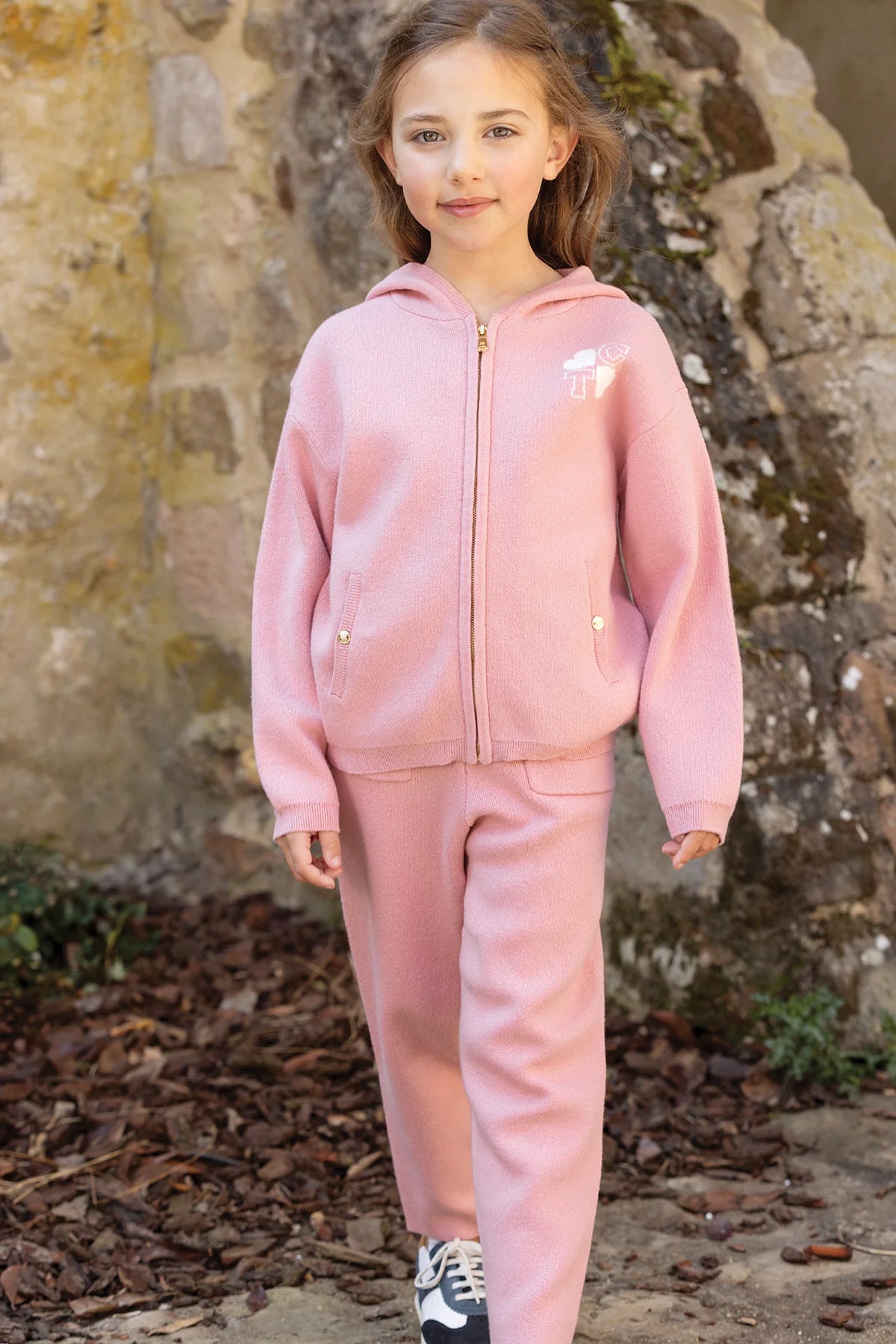 A young girl wearing the Tartine et Chocolat Girls’ Wool Hood Cotton Jogging Suit, featuring a zip-up hoodie and matching pants in pink, stands outdoors on a stone path with a stone wall in the background.