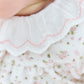 Close-up of a child wearing the Tartine et Chocolat Baby Girls’ Ruffle Collar Pajama, featuring a white outfit with a scalloped pink-edged collar and a floral pattern with green and pink accents.