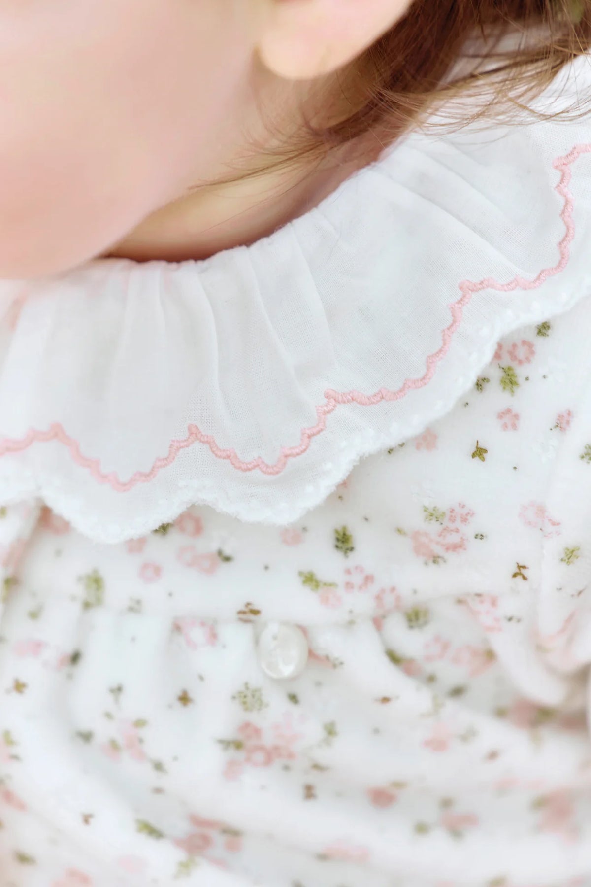 Close-up of a child wearing the Tartine et Chocolat Baby Girls’ Ruffle Collar Pajama, featuring a white outfit with a scalloped pink-edged collar and a floral pattern with green and pink accents.