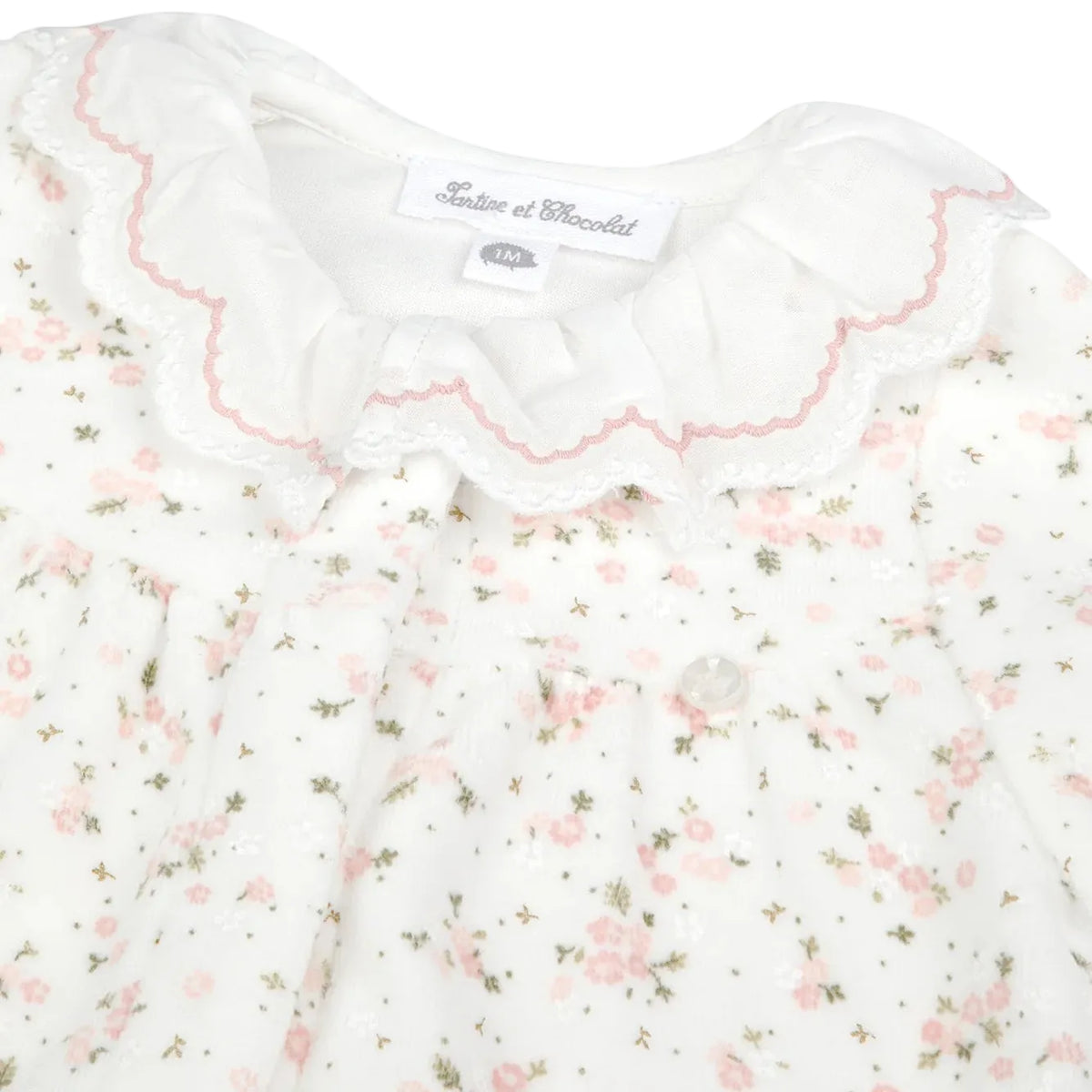 Introducing the Baby Girls’ Ruffle Collar Pajama by Tartine et Chocolat—a charming white baby pajama featuring a ruffled collar, scalloped lace trim, and delicate pink floral print. Expertly crafted with the same attention to detail as Tartine et Chocolat’s signature pajamas for girls, this elegant piece captures the timeless appeal of classic baby sleepwear. The label reads “Tartine et Chocolat.”.