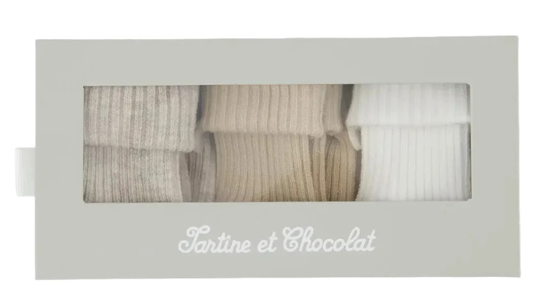 A rectangular box labeled “Tartine et Chocolat” in white script displays the Cotton Sock Set, featuring three pairs of ribbed toddler socks in gray, beige, and white. Made from gentle materials suitable for sensitive baby skin, the set is visible through a clear window on the box.