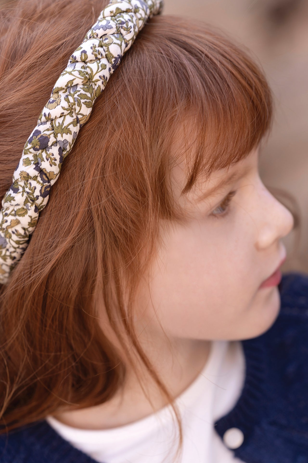 A young child with light skin and long brown hair is seen in profile, wearing the Tartine et Chocolat Liberty Print Braid Headband and a navy blue sweater, gazing off to the right.