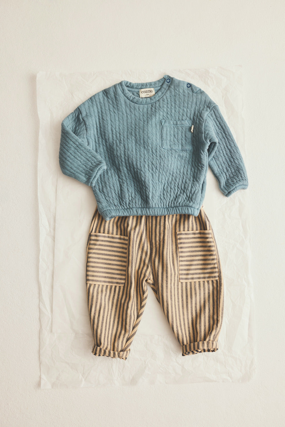 A Cozmo Baby Boys’ Chipre Quilted Sweatshirt in blue, featuring long sleeves and a chest pocket, paired with brown striped pants with patch pockets, both displayed flat on a white background.
