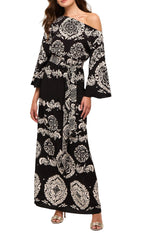 A woman stands wearing the Figue Christy Dress—a long black viscose crepe dress with white ornate patterns, a belted waist, off-the-shoulder neckline, and long sleeves—paired with silver heeled sandals.