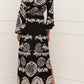 A woman stands in a corner facing away from the camera, wearing the Figue Christy Dress, a black and white patterned viscose crepe off-the-shoulder maxi with an elastic waistband, paired with white heeled sandals.