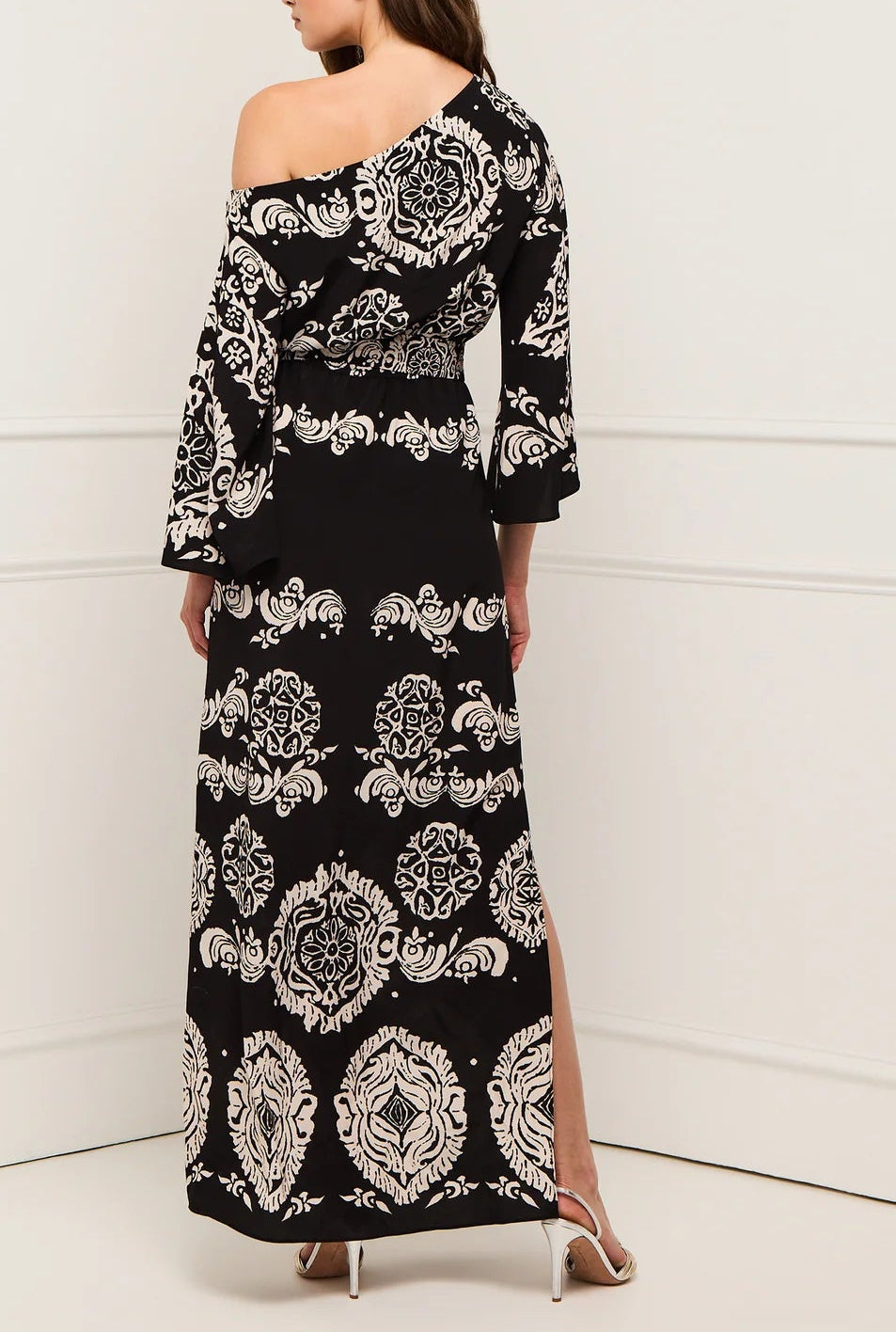 A woman stands in a corner facing away from the camera, wearing the Figue Christy Dress, a black and white patterned viscose crepe off-the-shoulder maxi with an elastic waistband, paired with white heeled sandals.