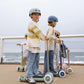 Four children wearing helmets ride Banwood Eco Scooters from the Eco Collection along a paved path by the ocean, with a fence and waves in the background.