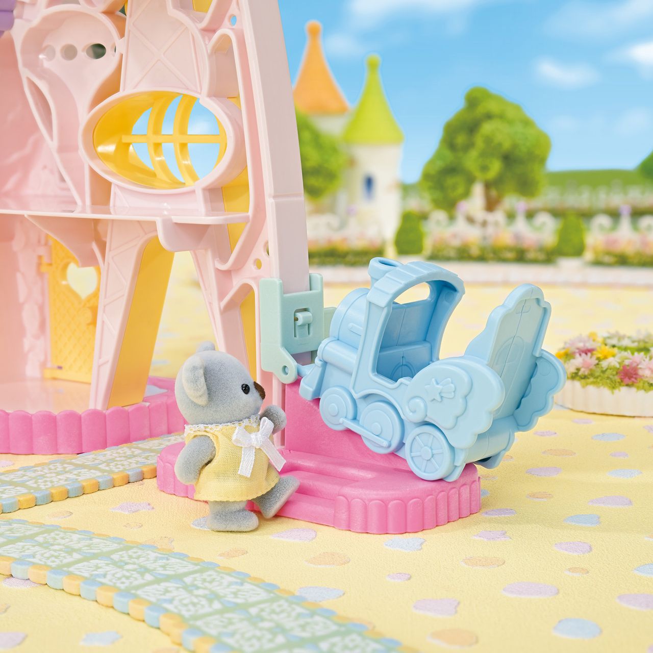 A small Calico Critters figure in a yellow dress stands near a blue toy train and the Baby Dream Amusement Park playset, which features pastel colors, a Ferris wheel, and castle elements, all set against a whimsical, colorful backdrop.