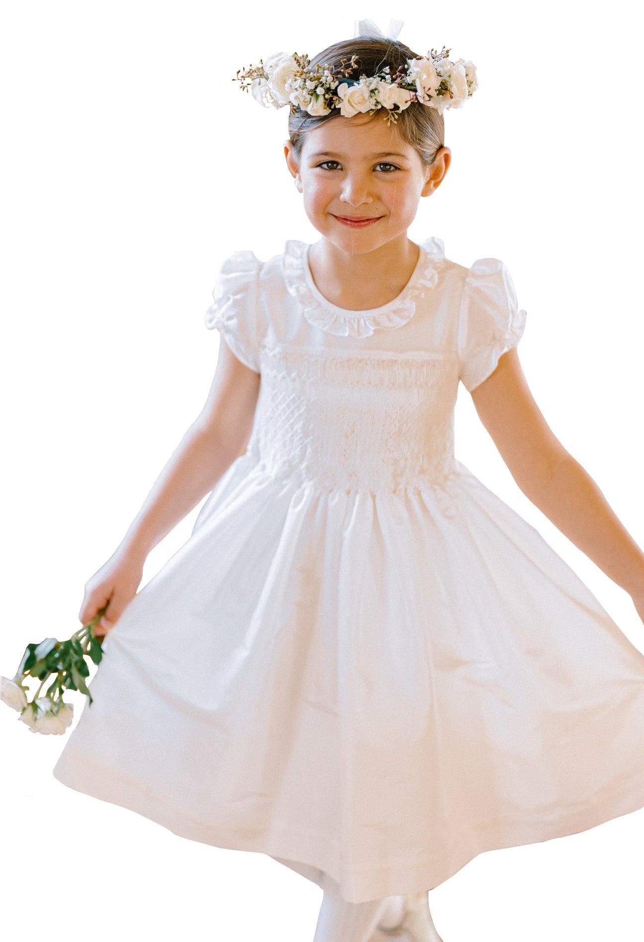 A young girl wearing the Antoinette Girls' Colombe White Silk Dress and a floral headband holds a small bouquet of flowers. She stands against a white background, smiling, making it perfect for special occasions.