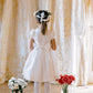 A young girl in an Antoinette Colombe White Silk Dress stands facing gold curtains, her hair tied with a white ribbon. Two vases, one with white flowers and the other with red flowers, are placed on the floor in front of her. This dress seems perfect for special occasions.