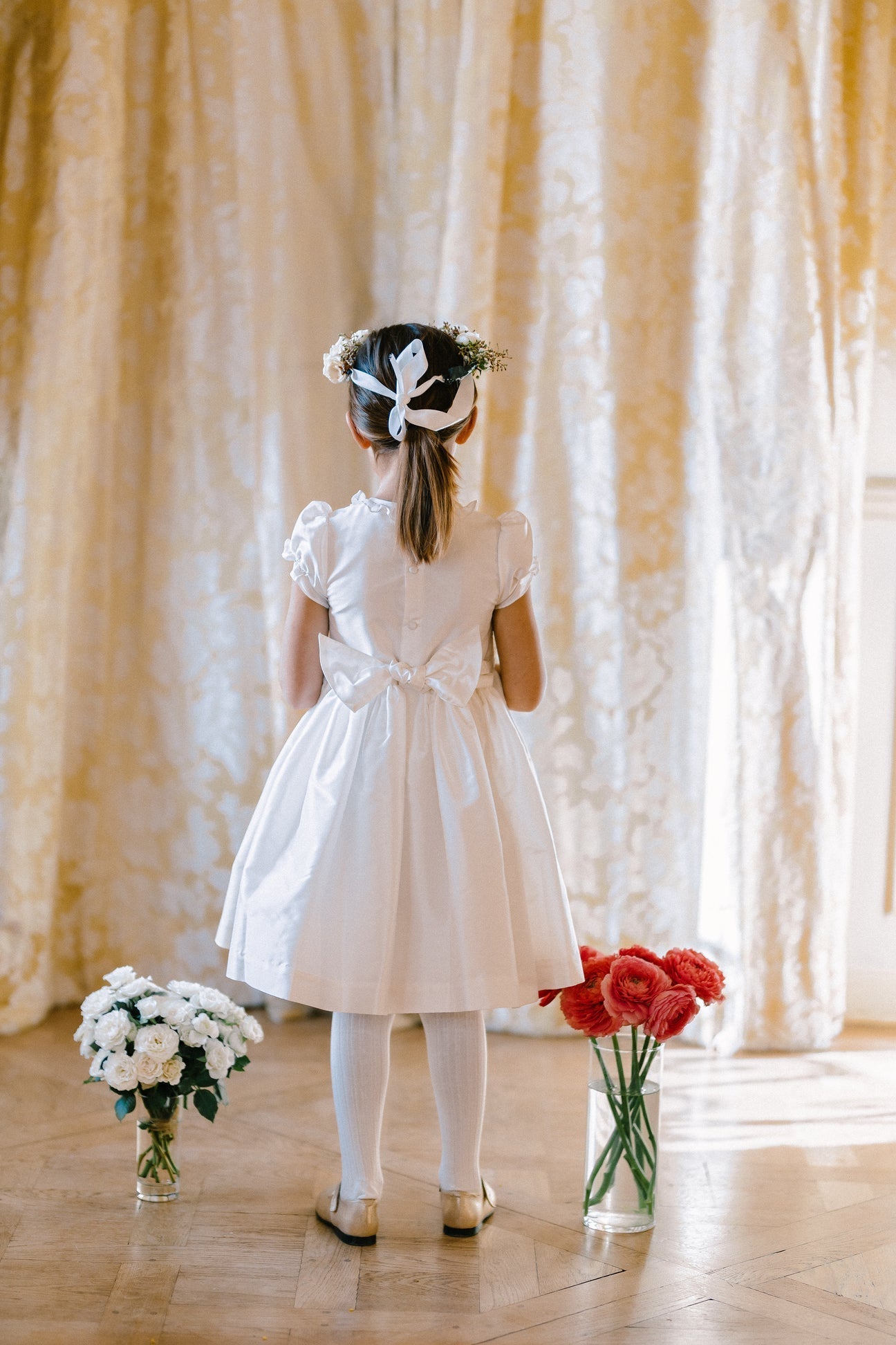 A young girl in an Antoinette Colombe White Silk Dress stands facing gold curtains, her hair tied with a white ribbon. Two vases, one with white flowers and the other with red flowers, are placed on the floor in front of her. This dress seems perfect for special occasions.