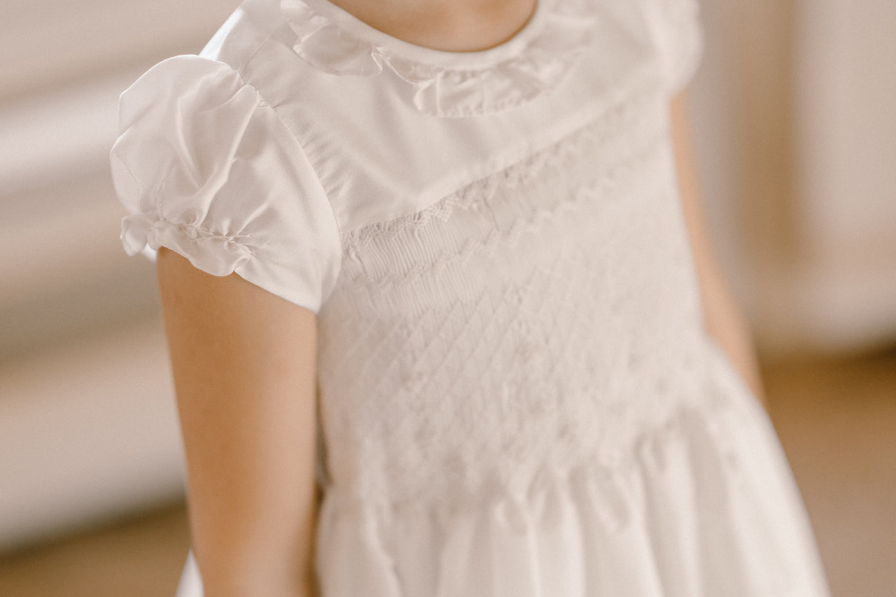 Child wearing the Antoinette Girls' Colombe White Silk Dress, featuring puff sleeves, a smocked bodice, and a frilled collar—perfect for special occasions.