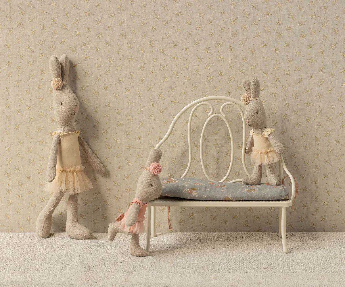 Three fabric bunny dolls in pale dresses are posed around the Maileg Vintage Bench, Miniature - Off White, set against a patterned wallpaper background.