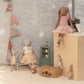 Five Maileg Bunny size 3 dolls in pastel outfits are gathered around wooden furniture; one, the Classic in Ballet suit and cream skirt, evokes a dancer. The softly lit scene includes a potted plant, tea set, and decorative suitcase.