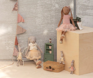 Five Maileg Bunny size 3 dolls in pastel outfits are gathered around wooden furniture; one, the Classic in Ballet suit and cream skirt, evokes a dancer. The softly lit scene includes a potted plant, tea set, and decorative suitcase.