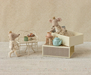 Maileg’s Royal Twin Mice, Little Sister & Brother in Matchbox, wear golden crowns and are shown by a table with tea and pastries, next to stacked matchbox beddings against a patterned wall.