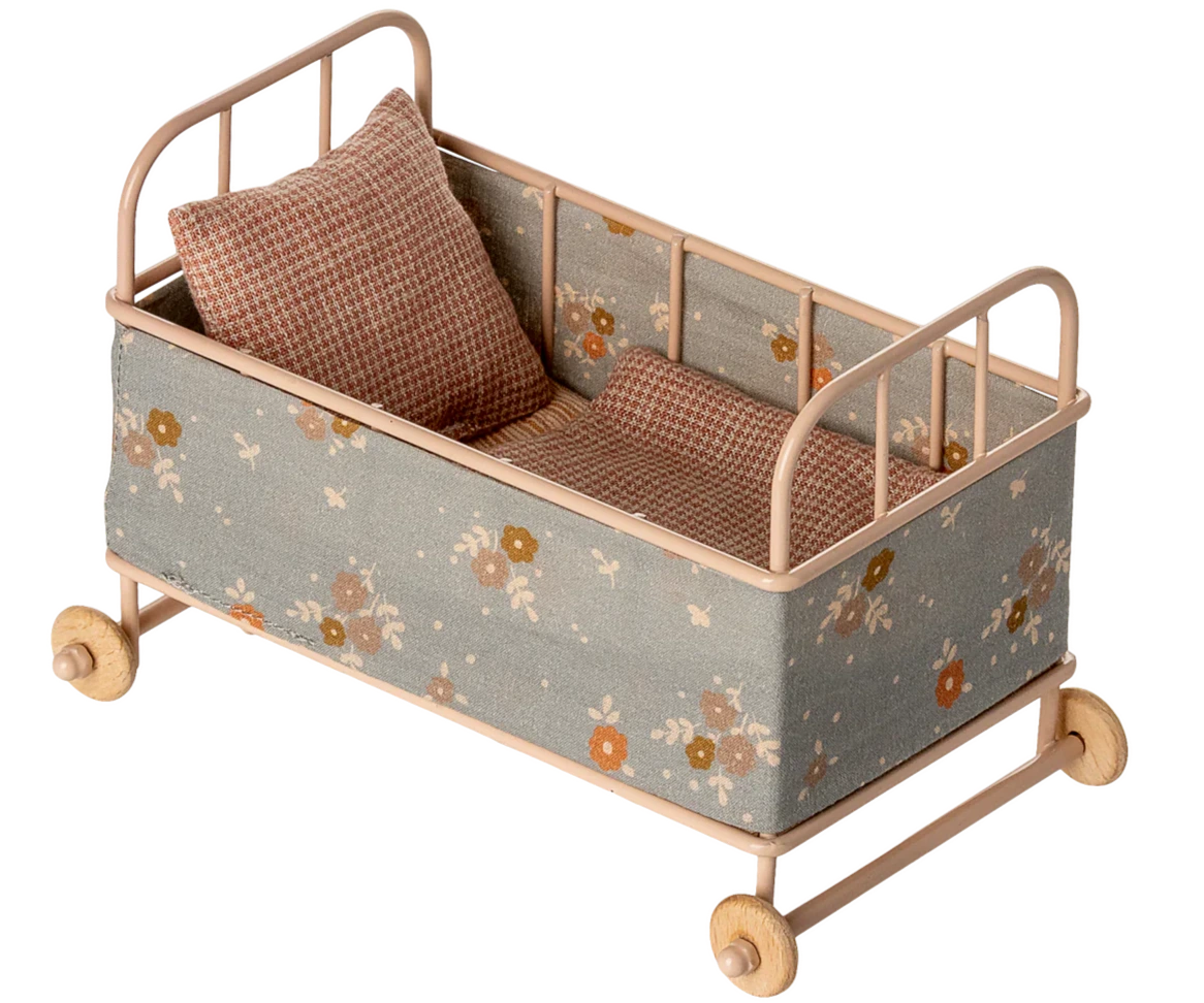 The Maileg Cot Bed, Micro – Powder is a small beige metal bed with floral fabric sides, wooden wheels, a soft mattress, and two checkered pillows—ideal for your micro sized friends.