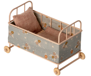 The Maileg Cot Bed, Micro – Powder is a small beige metal bed with floral fabric sides, wooden wheels, a soft mattress, and two checkered pillows—ideal for your micro sized friends.