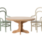 An oval wooden Maileg "Oval Dining Table, Mouse - Nature" has two curved-back chairs, one green and one cream, placed on either side against a plain white background.