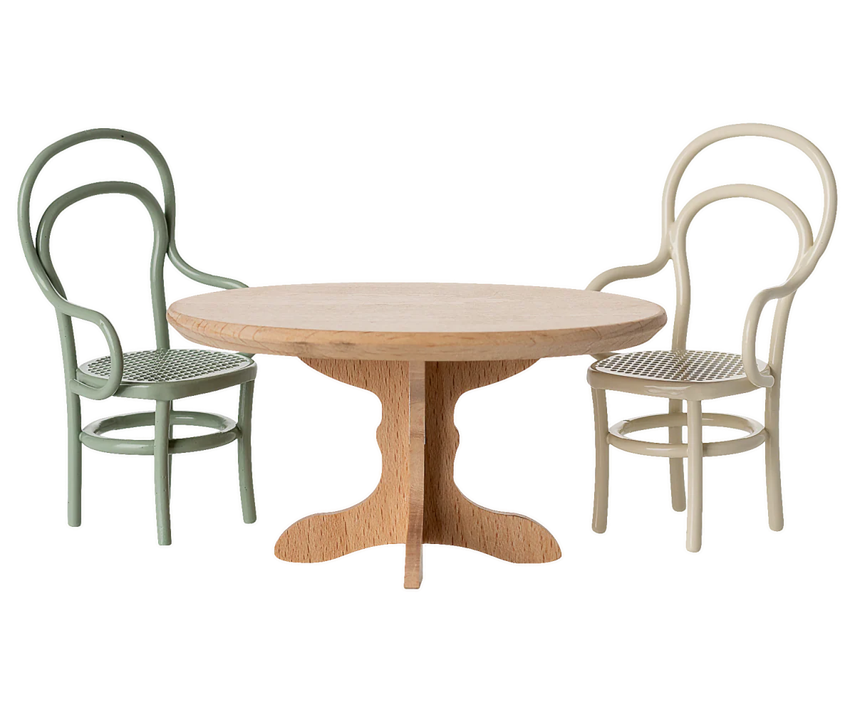 An oval wooden Maileg "Oval Dining Table, Mouse - Nature" has two curved-back chairs, one green and one cream, placed on either side against a plain white background.