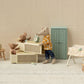 The Maileg Sleepy Wakey Baby Mouse in Matchbox - Rose lies in a cozy dollhouse scene with a dresser, clock, stool, green door, drying rack, and tiny décor on a textured surface against a dotted background.