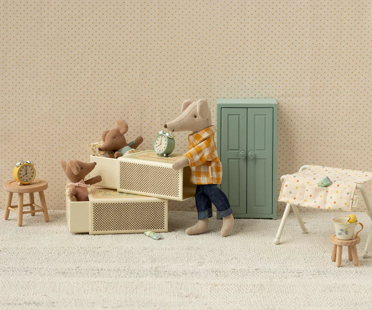 The Maileg Sleepy Wakey Baby Mouse in Matchbox - Rose lies in a cozy dollhouse scene with a dresser, clock, stool, green door, drying rack, and tiny décor on a textured surface against a dotted background.