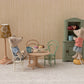 Two toy mice in a miniature room: one sits at a Maileg Oval Dining Table, Mouse – Nature set with mugs and pastries, while the other stands on a stool reaching into a green cupboard.
