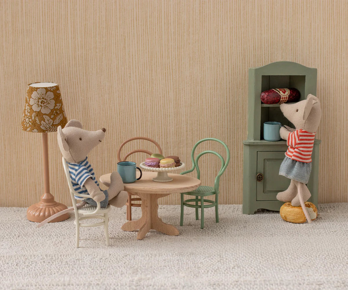 Two toy mice in a miniature room: one sits at a Maileg Oval Dining Table, Mouse – Nature set with mugs and pastries, while the other stands on a stool reaching into a green cupboard.