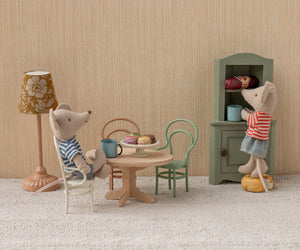 Two toy mice in a miniature room: one sits at a Maileg Oval Dining Table, Mouse – Nature set with mugs and pastries, while the other stands on a stool reaching into a green cupboard.