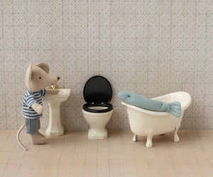 A Maileg Off White Water Closet - Mouse stands by a sink next to a compact toilet, with a plush fish resting in a small bathtub—all arranged in a charming miniature bathroom scene.