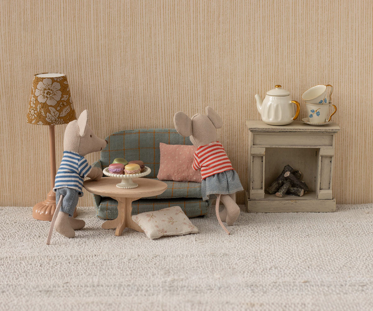 Two toy mice in striped shirts are relaxing in their cozy mouse home, complete with a Mouse Sofa Bed, a Maileg Nature Oval Coffee Table – Mouse topped with desserts, a lamp, and a fireplace adorned with a teapot and cups.
