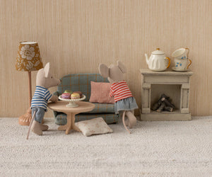 Two toy mice in striped shirts are relaxing in their cozy mouse home, complete with a Mouse Sofa Bed, a Maileg Nature Oval Coffee Table – Mouse topped with desserts, a lamp, and a fireplace adorned with a teapot and cups.