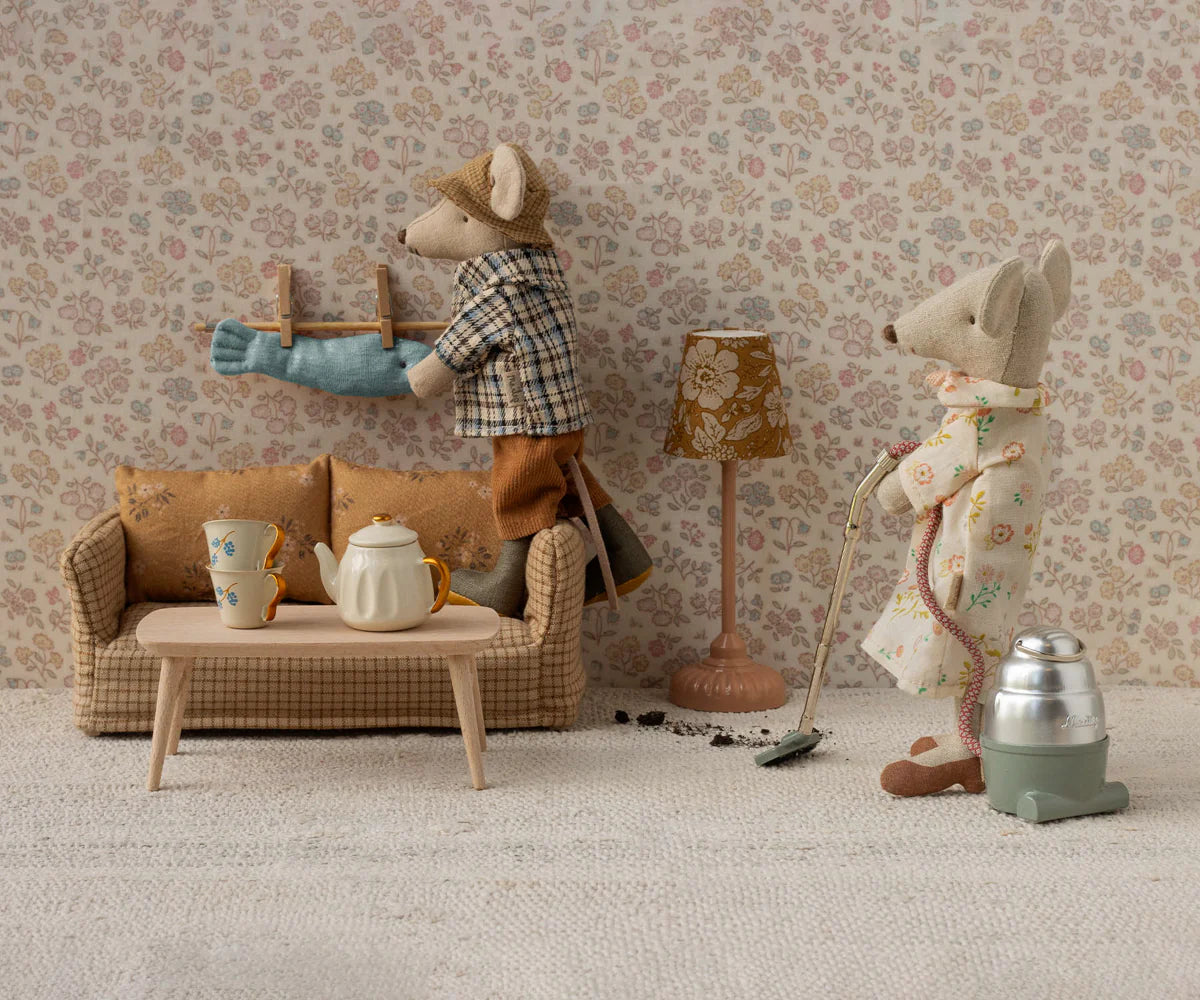 Two toy mice from Maileg tidy their miniature living room; one vacuums while the other hangs laundry. The cozy scene features the Maileg Couch, Mouse - Mable, a table with tea set, and a lamp.