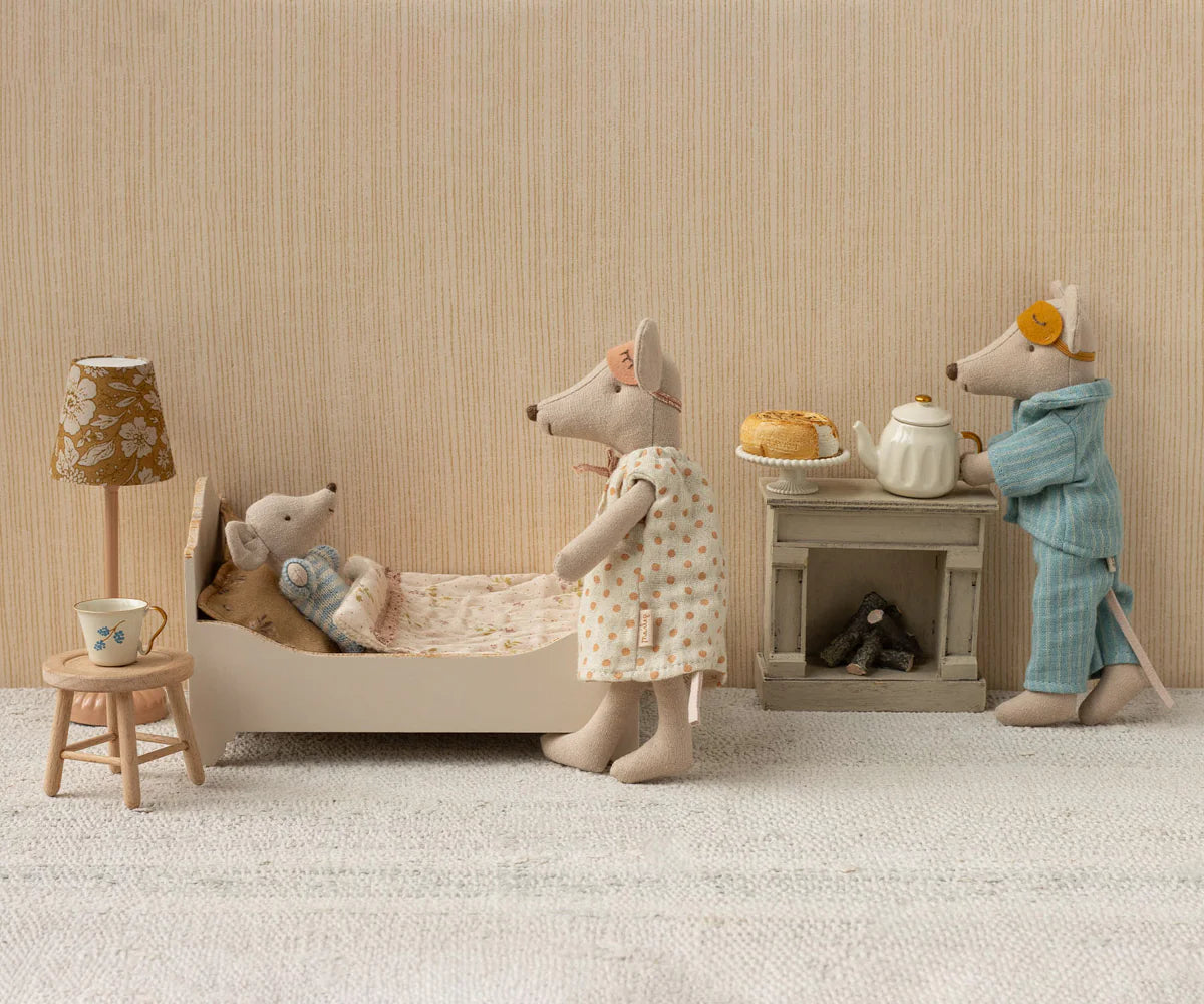 Three Maileg mice in a miniature bedroom: one snuggled in the Maileg Bed, Mouse - Off White, one standing nearby, and another in pajamas making tea and snacks on a tiny stove.
